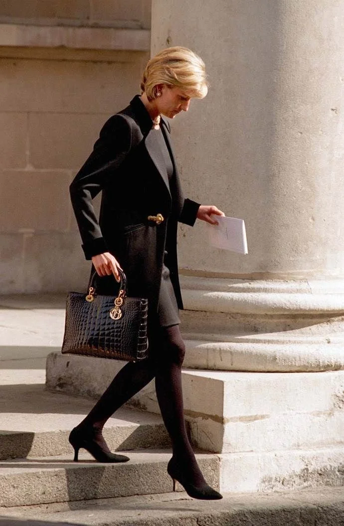 Princess Diana wearing a tailored black blazer and trousers, embodying the quiet money aesthetic with a structured handbag while walking outdoors.
