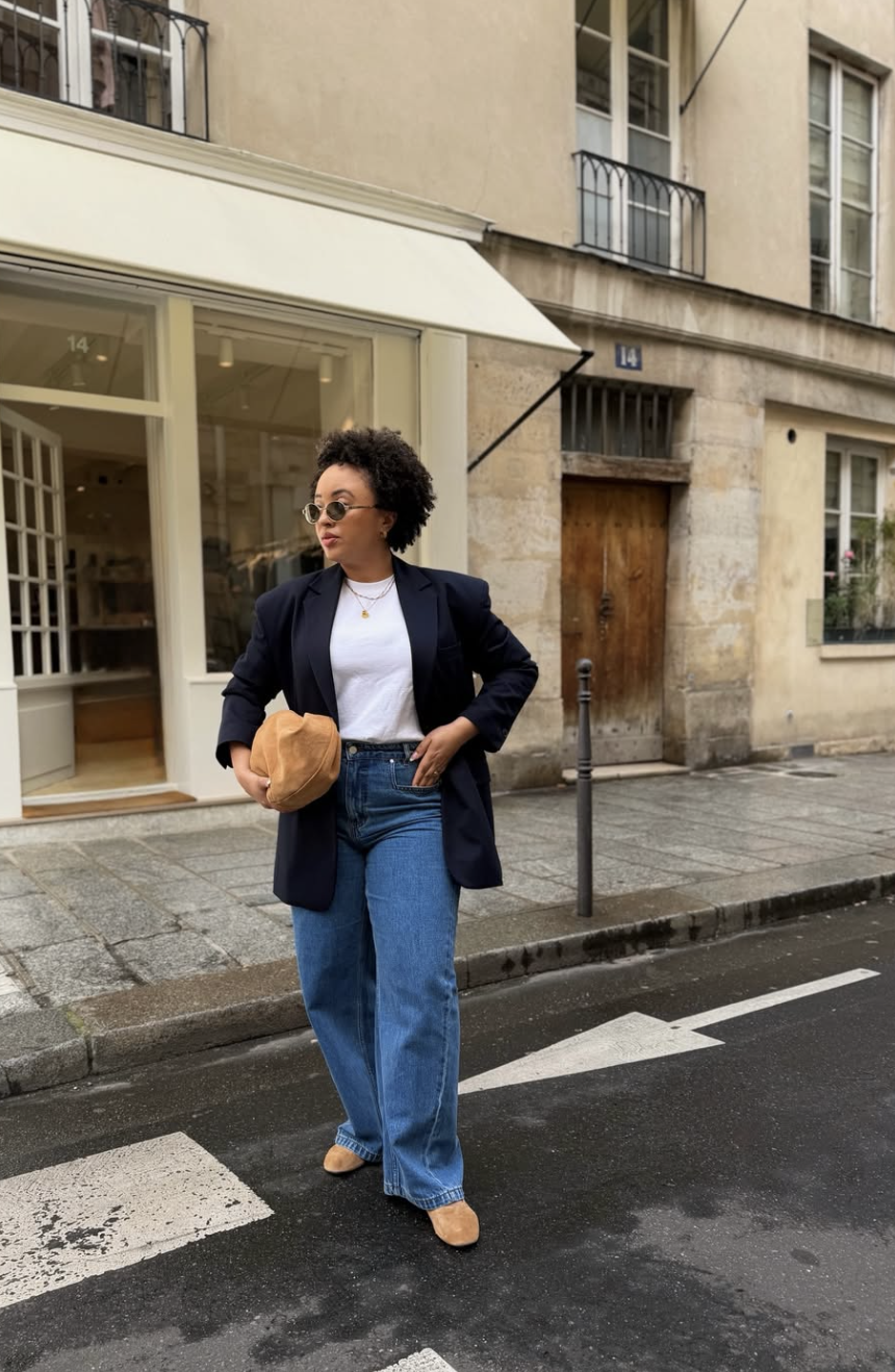 A woman walking on a city street wearing an oversized navy blue blazer over a white t-shirt, paired with medium-wash wide-leg jeans, tan suede shoes, and a brown clutch.