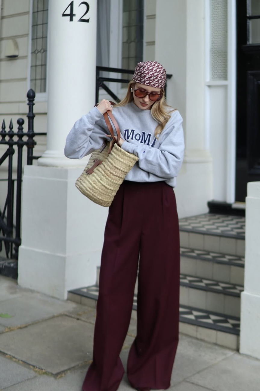 A close-up shot of a woman with long wavy blonde hair wearing a pink and white monogrammed silk scarf styled as a classic bandana tie, paired with a grey 'MoMA' sweatshirt, orange-tinted aviator sunglasses, and a woven straw tote bag.
