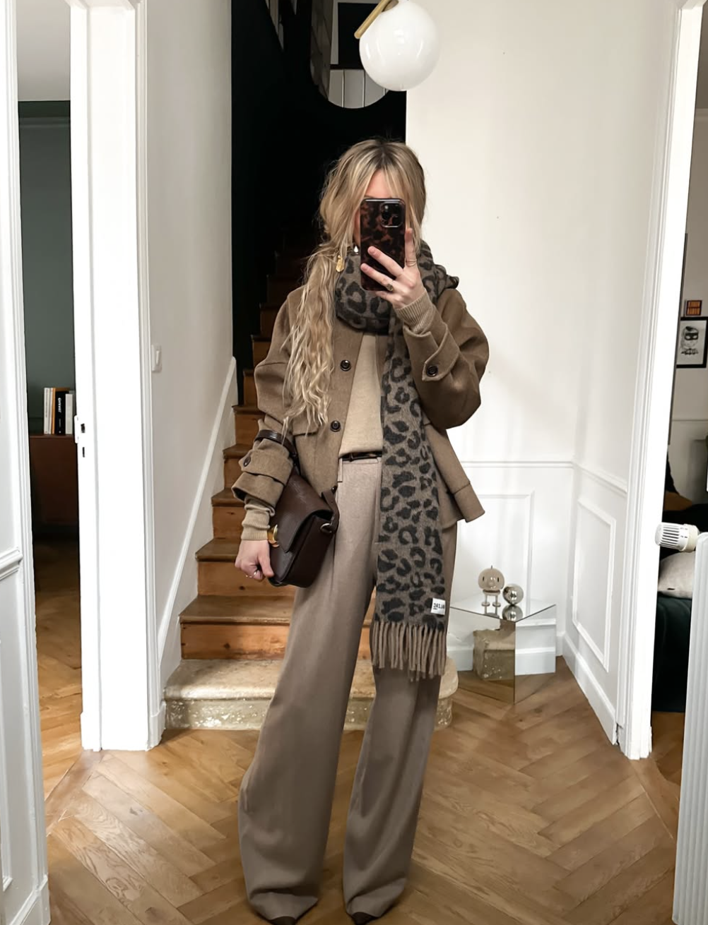 A woman wearing a tailored camel wool coat, tan trousers, and a long leopard print scarf, carrying a dark brown leather shoulder bag.