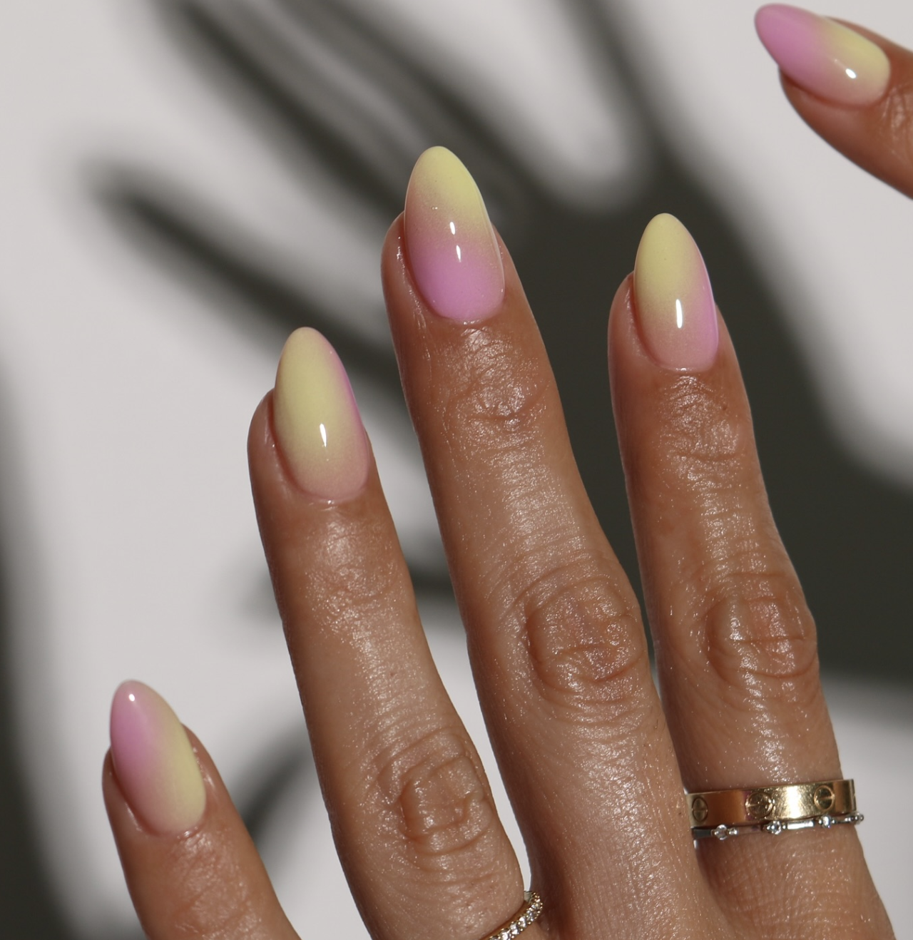 A close-up of long, almond-shaped nails featuring a soft, pastel aura gradient of pink and yellow, heavily accessorized with stacked gold rings.