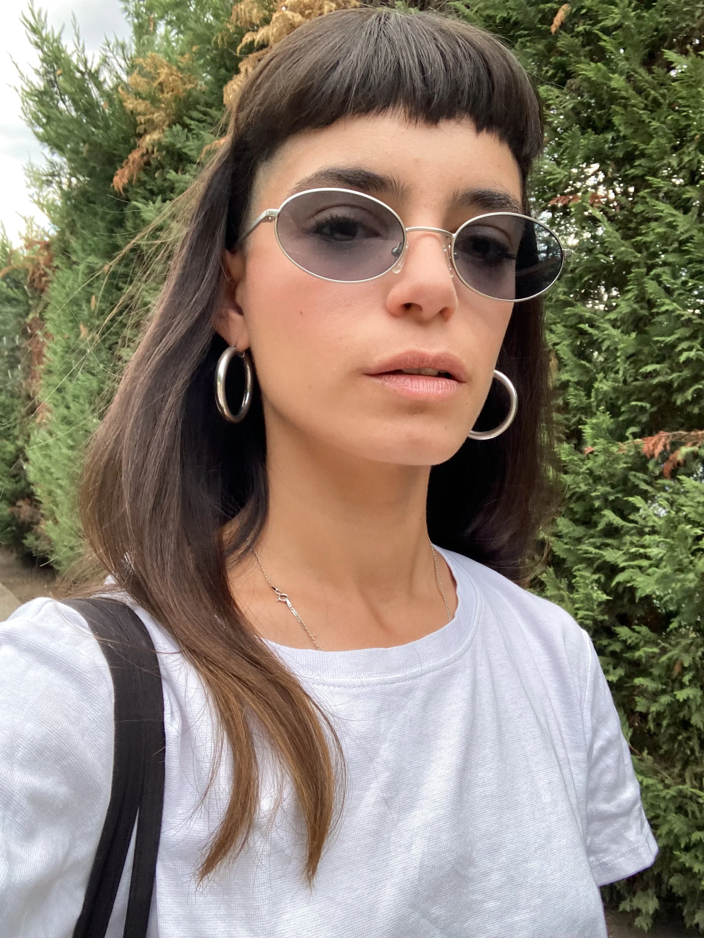 A portrait of Sofia Sencion wearing silver tinted oval sunglasses, styled with a white t-shirt and large silver hoop earrings against a natural green background.