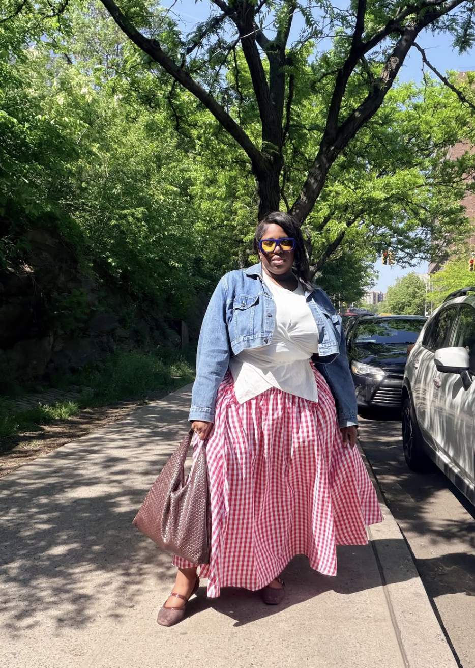 Plus-size woman wearing a red and white gingham A-line maxi skirt paired with a white tee, cropped denim jacket, and a woven brown tote bag.