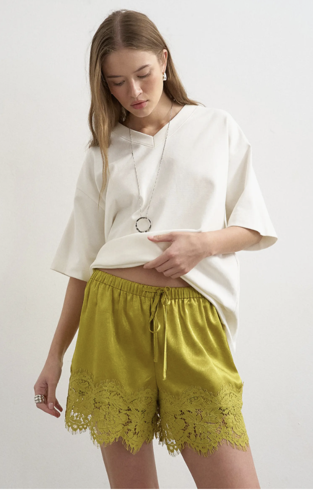 Close-up of lime green chartreuse satin shorts with an elastic drawstring waist and heavy matching lace trim at the hem, paired with a relaxed white tee.