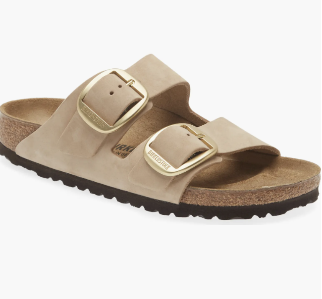 A studio product shot of a tan suede Birkenstock Arizona slide sandal featuring two wide adjustable straps with oversized gold buckles and a classic cork footbed.