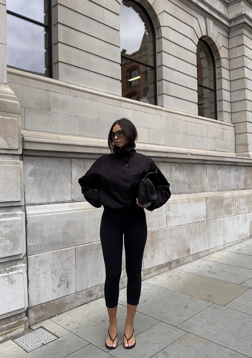 A woman wearing a black cropped puffer jacket with an oversized collar, high-waisted black leggings, and tan leather flip-flops, standing in front of a stone building with arched windows.