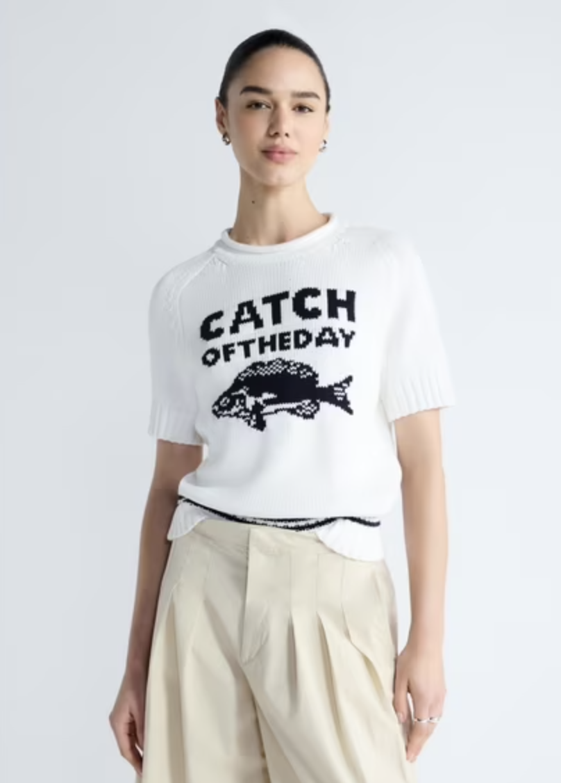 A woman styling a short-sleeve cream knit sweater featuring black "CATCH OF THE DAY" text and a fish graphic, paired with high-waisted pleated beige trousers.