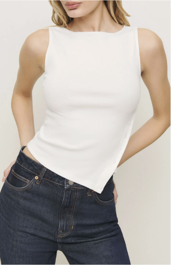 A close-up shot of a model wearing a ribbed cream-white knit tank top with an asymmetrical angled hem, paired with dark-wash blue jeans.