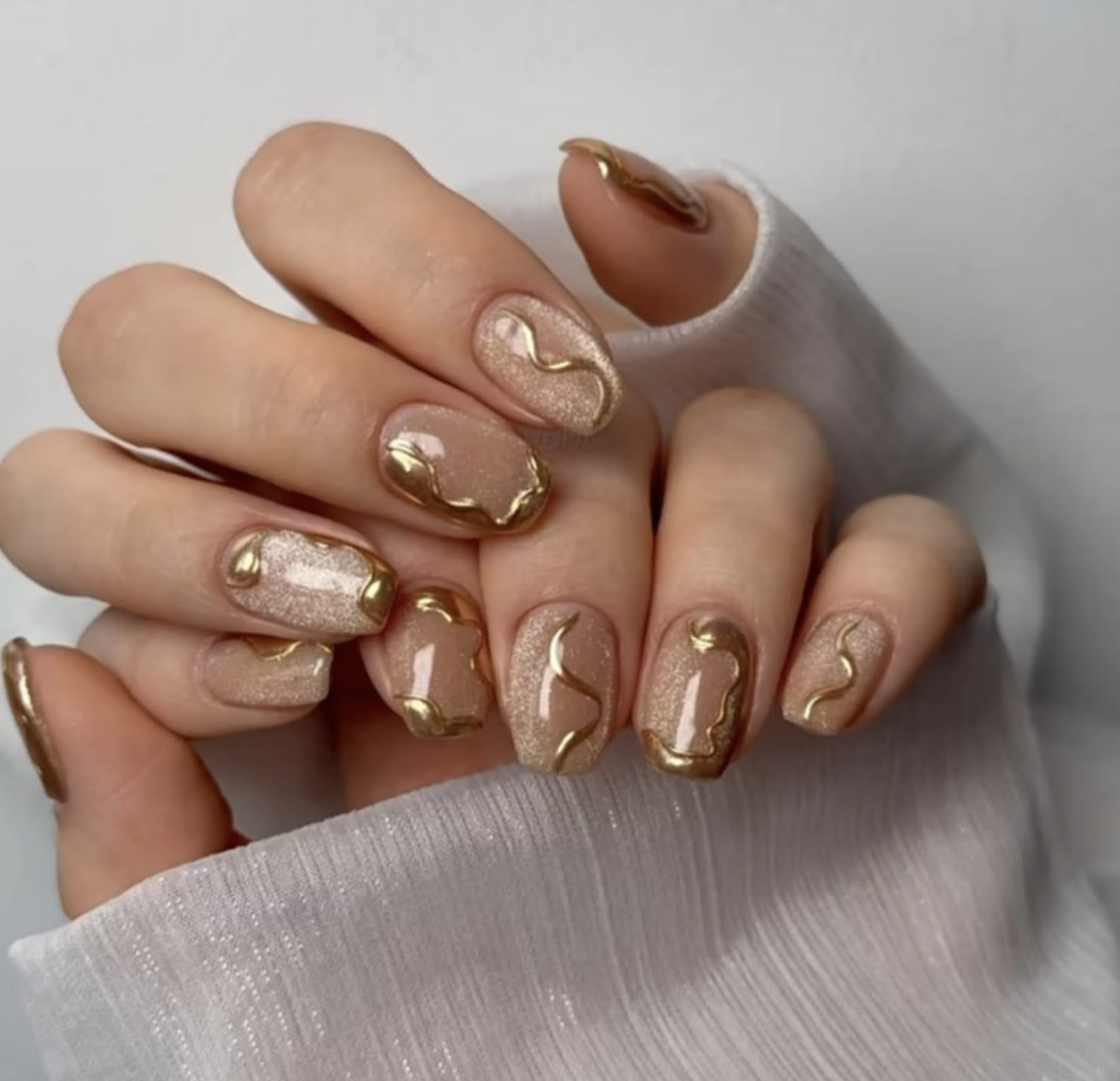 Short square press-on nails with a nude pink base, featuring a diagonal gold glitter stripe and fine golden line accents.
