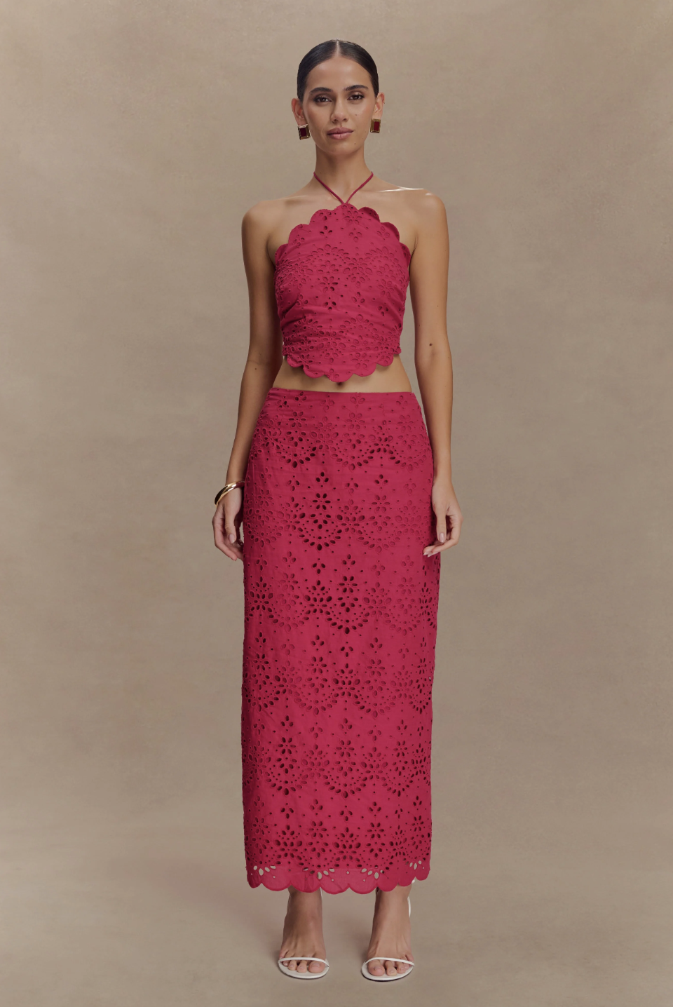 A woman wearing a vibrant magenta pink two-piece eyelet broderie set featuring a scalloped halter crop top and a matching high-waisted midi skirt, paired with white strappy heeled sandals.