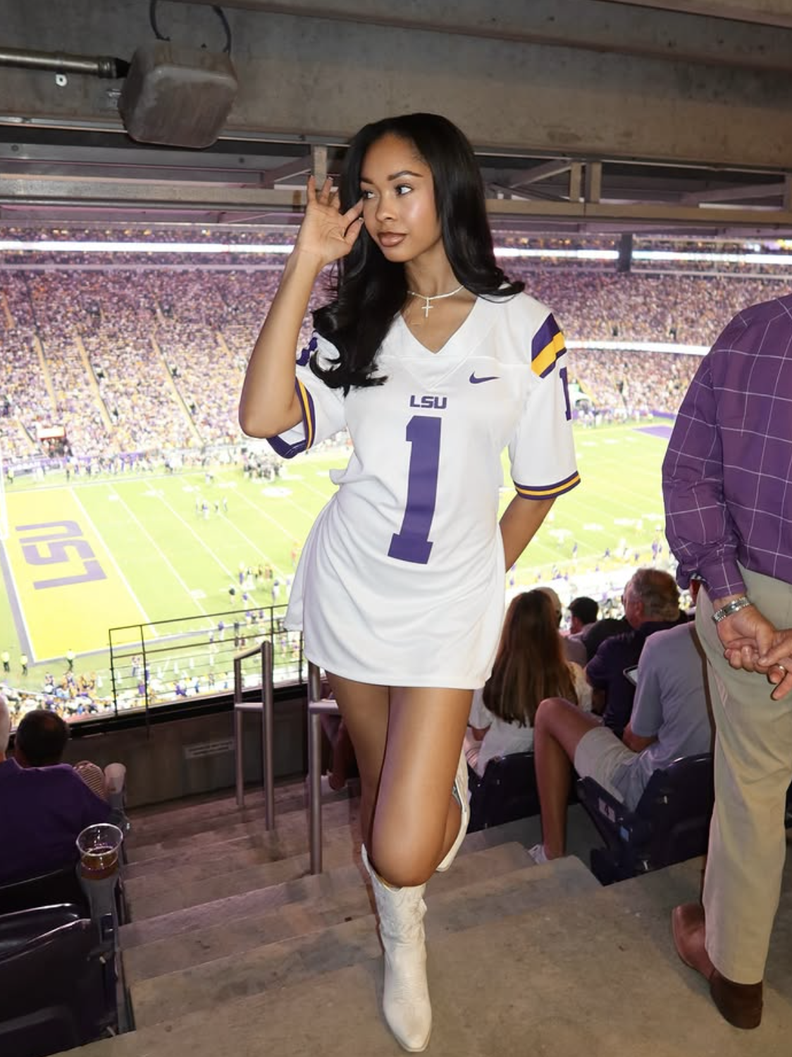 t: Woman in a football stadium suite wearing an oversized white LSU jersey as a mini dress with white cowboy boots.