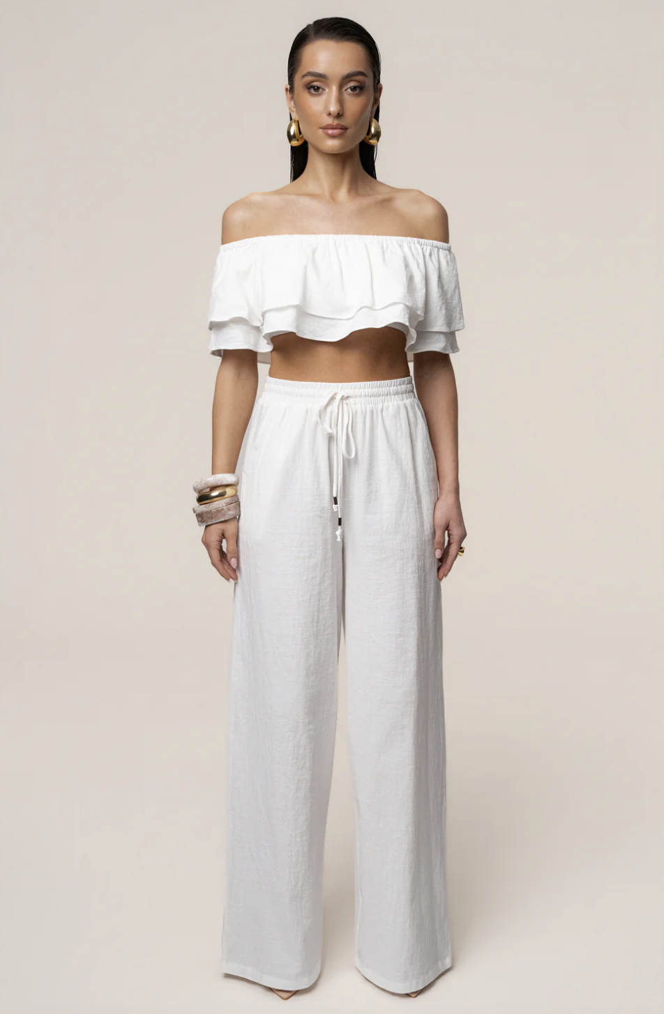 A woman wearing the JLUXLABEL White Marina Two-Piece Set featuring an off-the-shoulder ruffle top and matching wide-leg linen-style pants for a luxury Mediterranean vacation.
