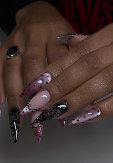 Close-up of a hand with long, stiletto-shaped nails featuring a dramatic summer manicure with a sheer pink base, bold black polka dots, and intricate black lace nail art accents.