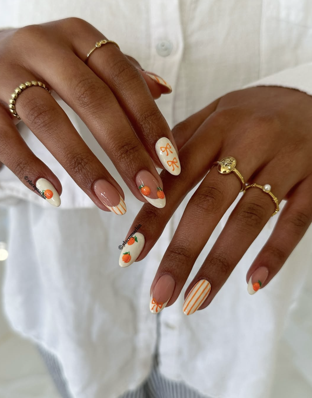 A close-up of almond-shaped nails featuring a creamy white and orange-striped French manicure, detailed with hand-painted mini oranges and vibrant orange bow accents, accessorized with fine gold rings.