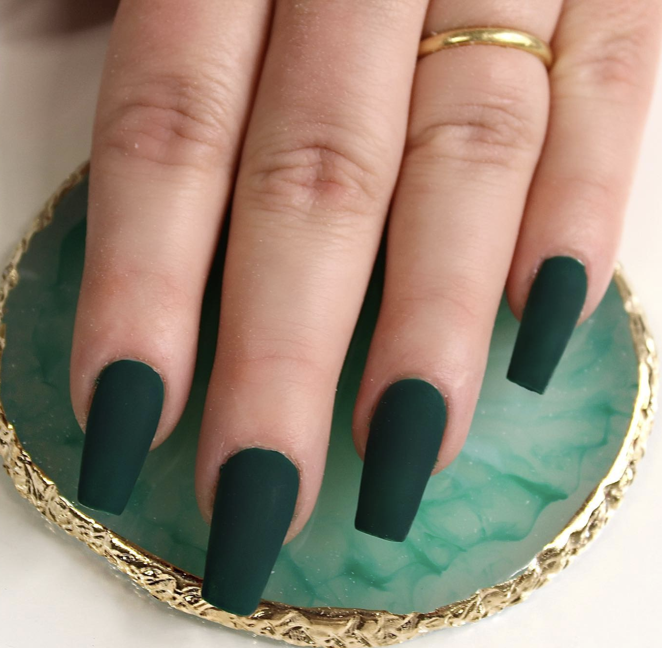 Long square-shaped gel nails with a deep forest green matte finish, shown over a green marble coaster.