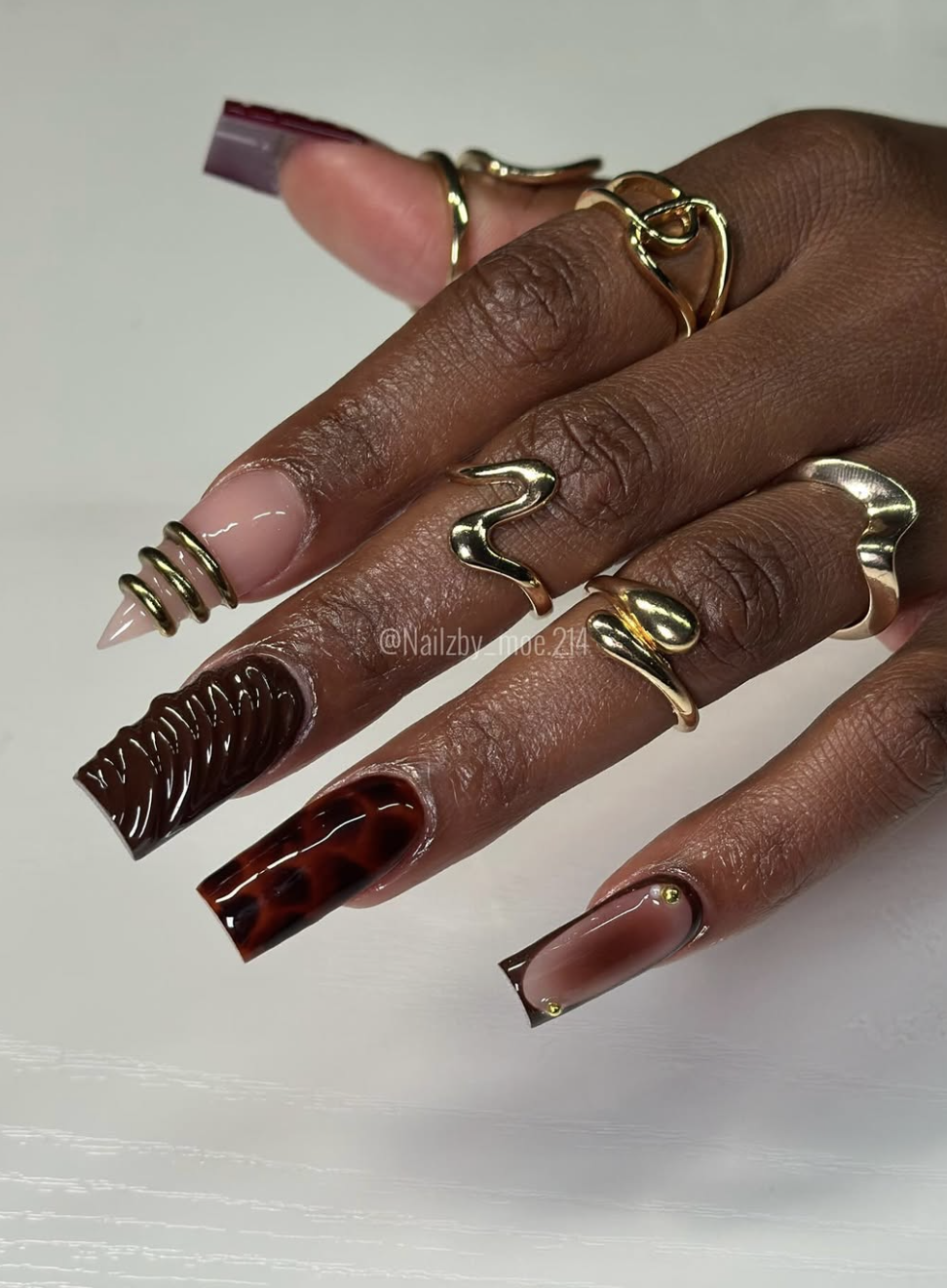 A close-up of long coffin-shaped nails featuring a mix of deep chocolate brown polish, gold wire cuffs, tortoiseshell pattern, and 3D textured swirl accents styled with gold rings.