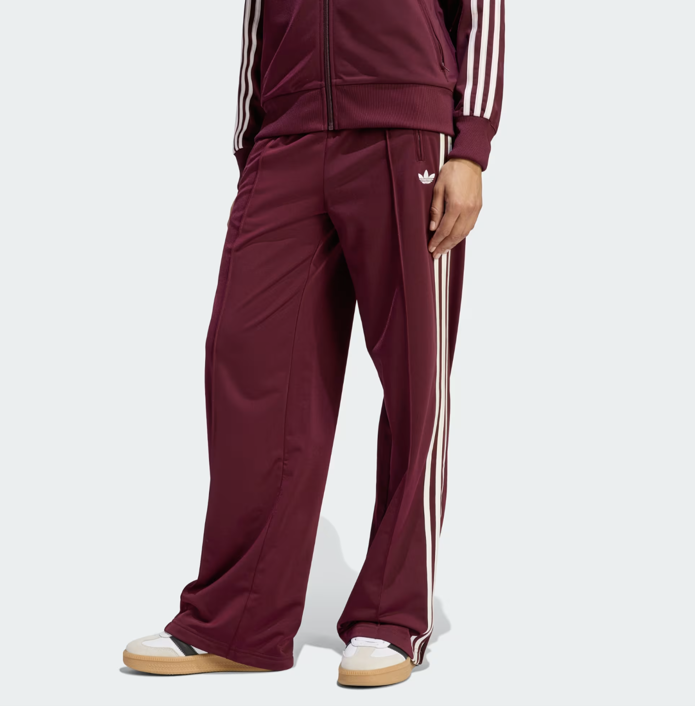 A close-up of the Adidas Firebird Loose Track Pants in a rich burgundy maroon with white side stripes, showing the high-waisted fit and embroidered Trefoil logo for a Sportif Luxe spring outfit.