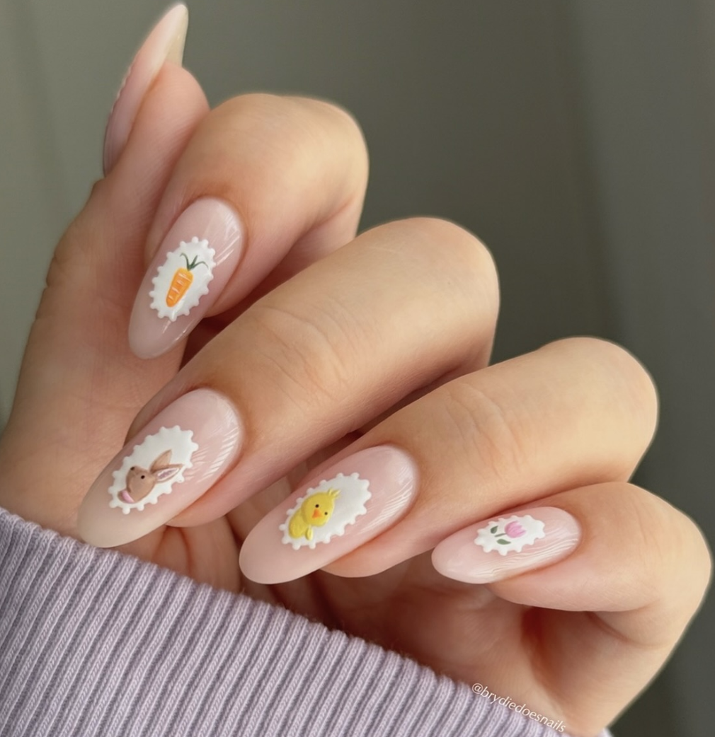 A close-up of almond-shaped nails with a milky white base featuring hand-painted vintage postage stamp designs with scalloped borders, including illustrations of a carrot, a bunny, a baby chick, and a flower.