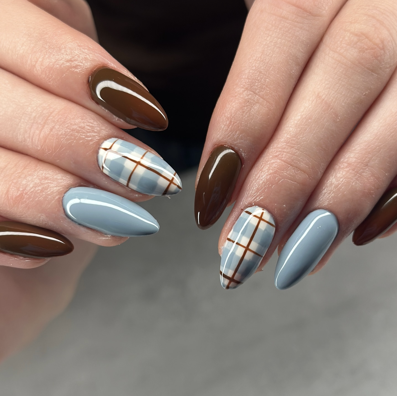 A close-up of almond-shaped nails featuring a mix of solid chocolate brown polish, light dusty blue, and a blue-and-white plaid print with brown grid lines.