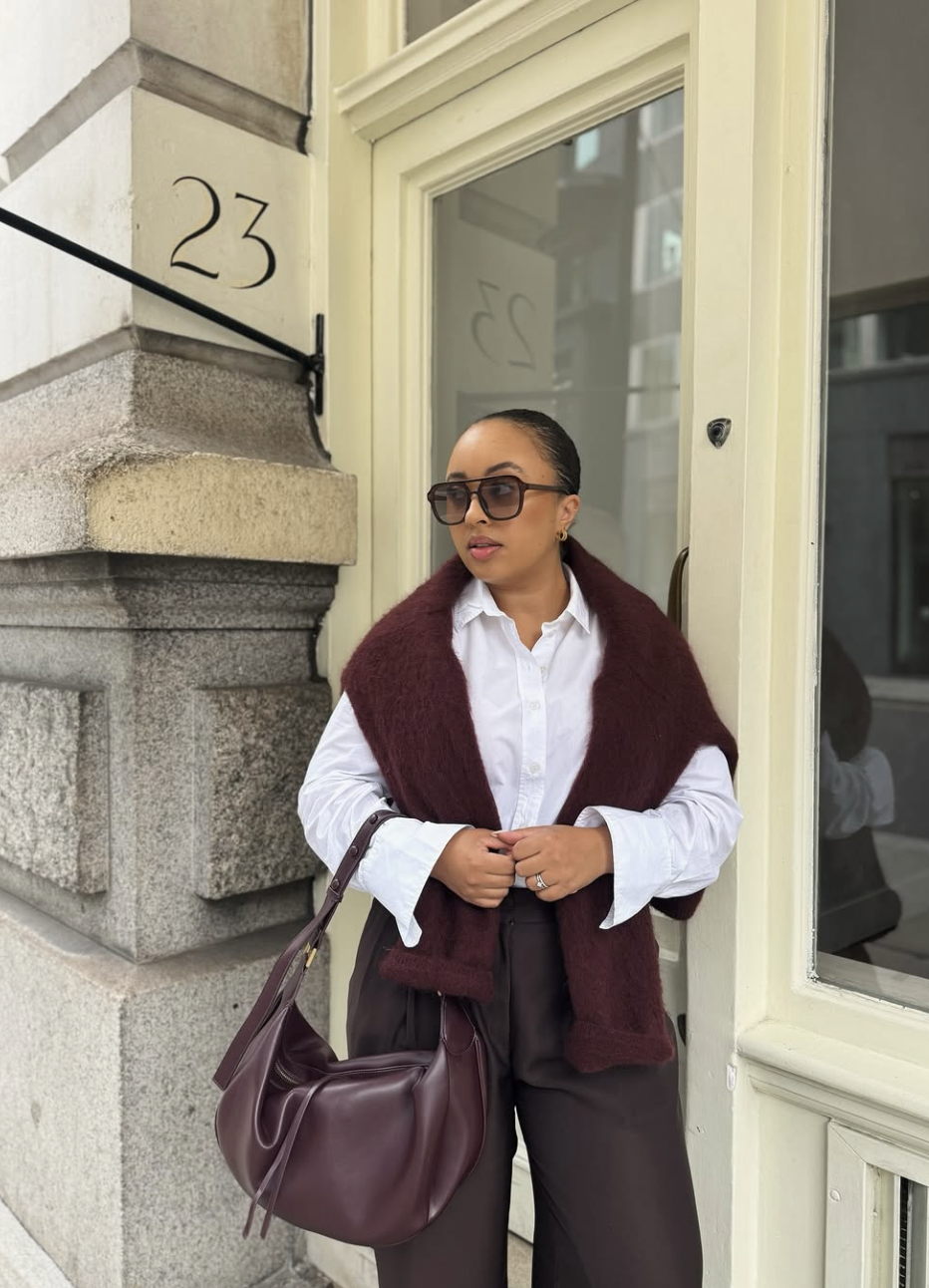 A woman wearing dark brown trousers with a white button-down shirt and a burgundy sweater draped over her shoulders, a chic brown pants outfit idea.