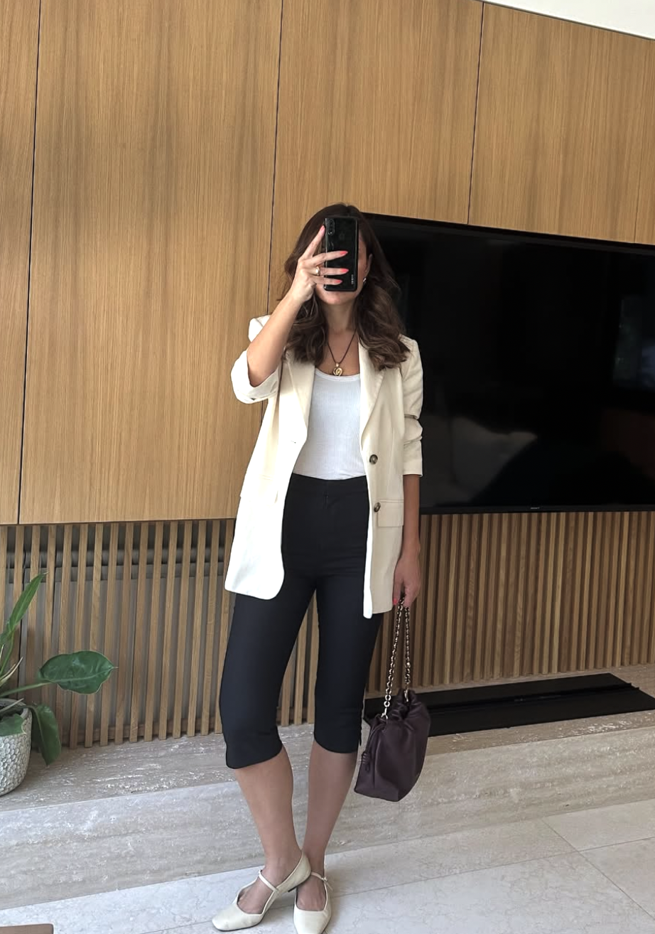 A fashion blogger taking a mirror selfie wearing an oversized white blazer, a white tank top, black capri pants, and black ballet flats.