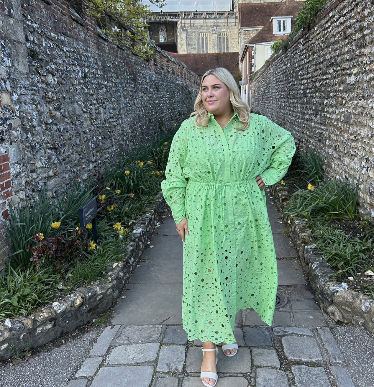 Plus-size woman wearing a vibrant lime green eyelet maxi dress with long sleeves and a collared neckline, standing on a cobblestone pathway.