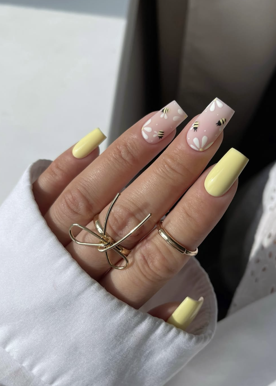 Long almond-shaped nails in a soft buttery yellow, featuring a white sunflower accent nail with small hand-painted honey bees and black flight trail details.