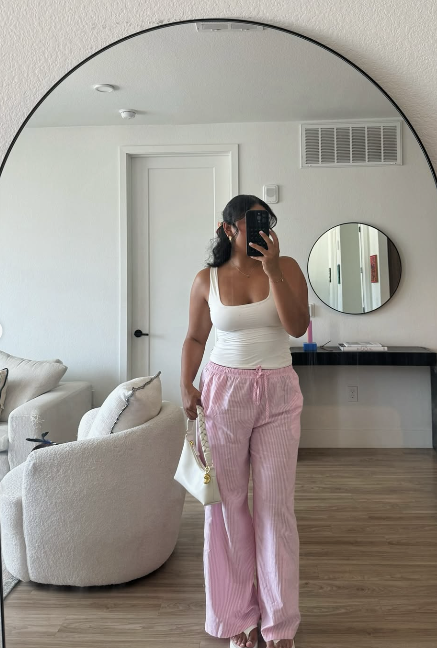 A woman taking a mirror selfie wearing a white ribbed tank top and high-waisted pink wide-leg linen pants in a modern neutral-toned room.