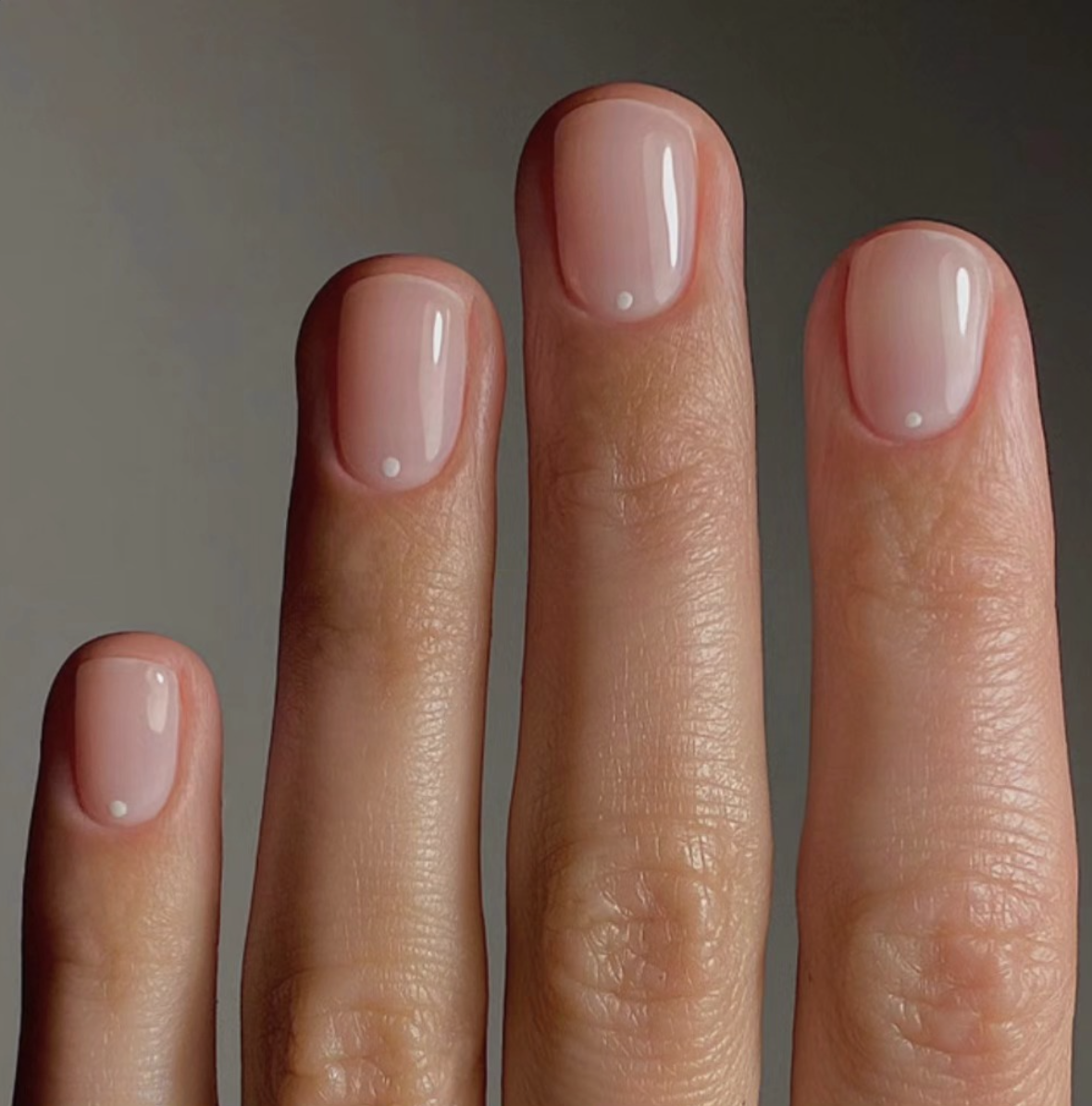 Short square nails with a sheer nude gloss and a single white dot detail at the base of each nail.