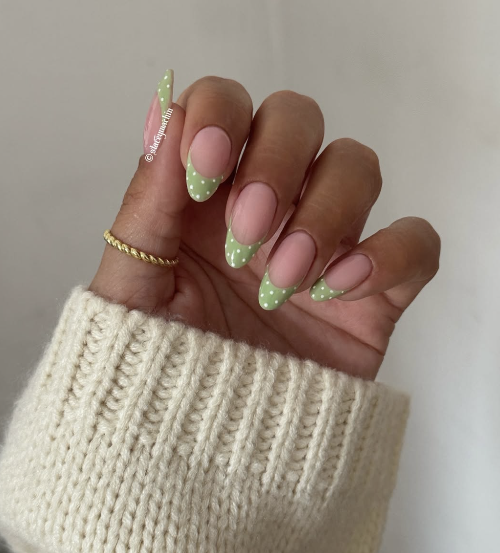 A close-up of almond-shaped nails featuring sheer nude polish with mint green french tips and white polka dot accents.