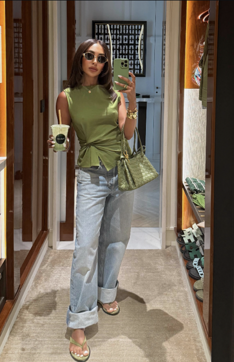 A woman wearing an olive green sleeveless peplum-style top with a tie-waist detail, styled with light wash wide-leg jeans and a matching green woven handbag.