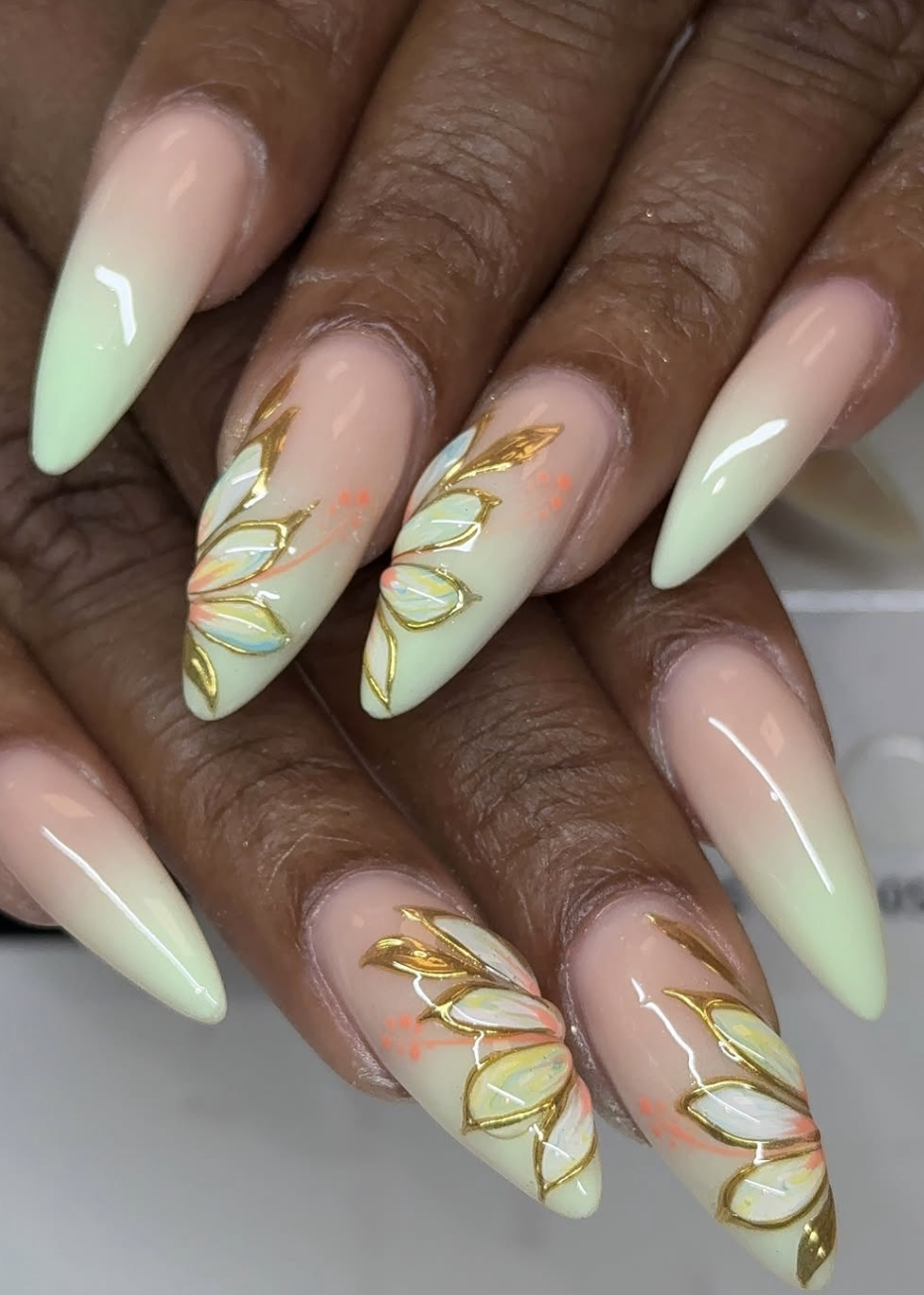 A close-up of almond-shaped nails featuring a mix of solid mint green and nude polish with gold metallic leaf art and abstract floral designs.