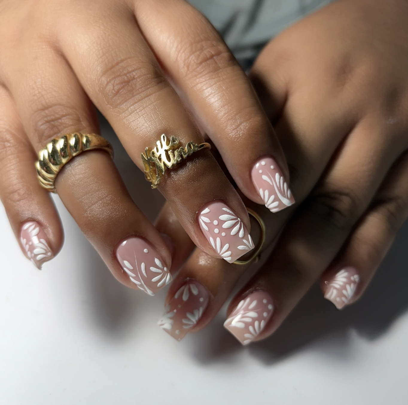 A close-up of almond nails with a sheer brown base featuring delicate white floral nail art, styled with gold statement rings.