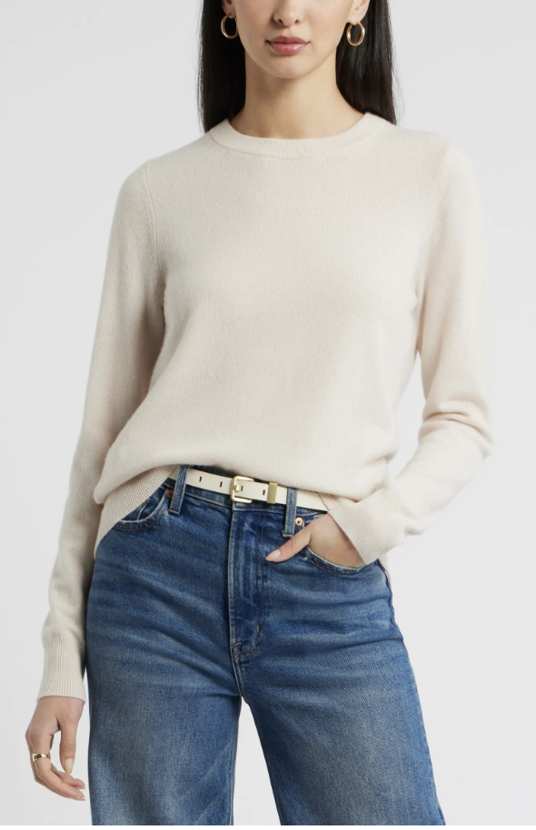 A close-up shot of a model wearing a plush oatmeal-colored cashmere crewneck sweater, tucked into high-waisted blue wide-leg jeans.