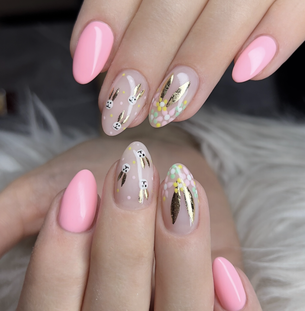 A close-up of almond-shaped nails featuring a mix of solid bubblegum pink and clear-base designs with gold foil bunny ears, metallic accents, and colorful "mini egg" speckles in mint, yellow, and white.