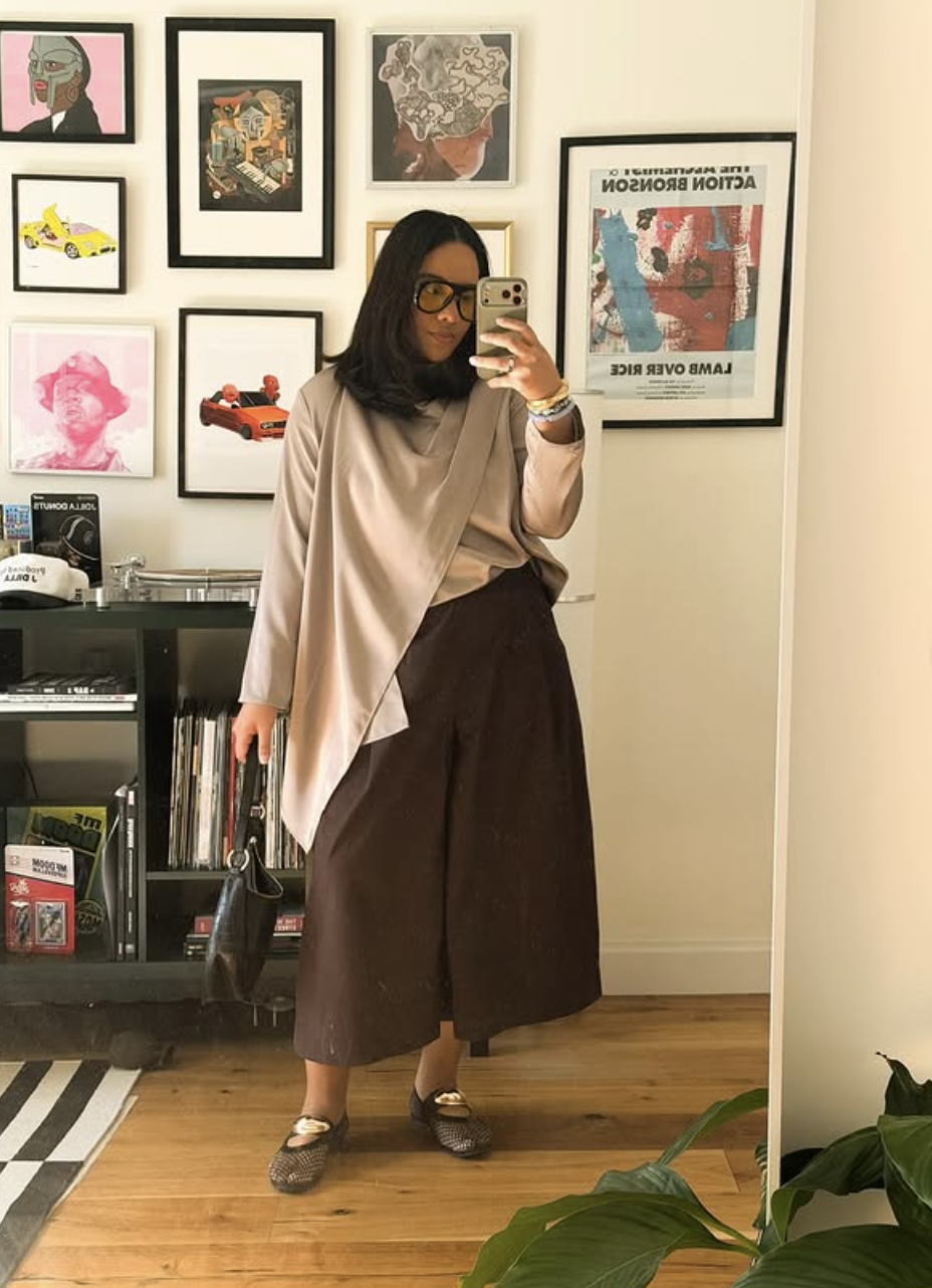 A mirror selfie of a woman wearing loose, structured dark brown Bermuda shorts, an asymmetrical oversized beige top, black-rimmed glasses, and metallic bronze jelly flat shoes in front of a gallery wall.