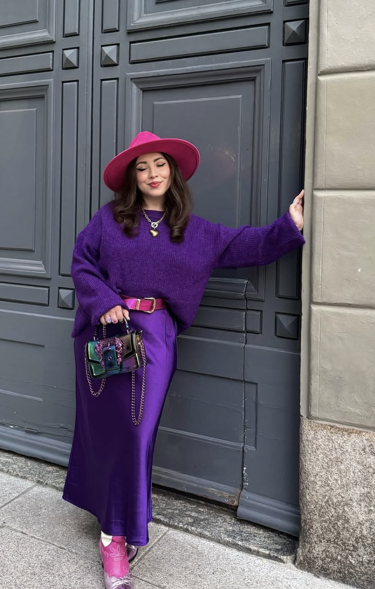 A woman wearing a monochromatic purple outfit featuring an oversized knit sweater, trousers, and a vibrant pink wide-brim hat.