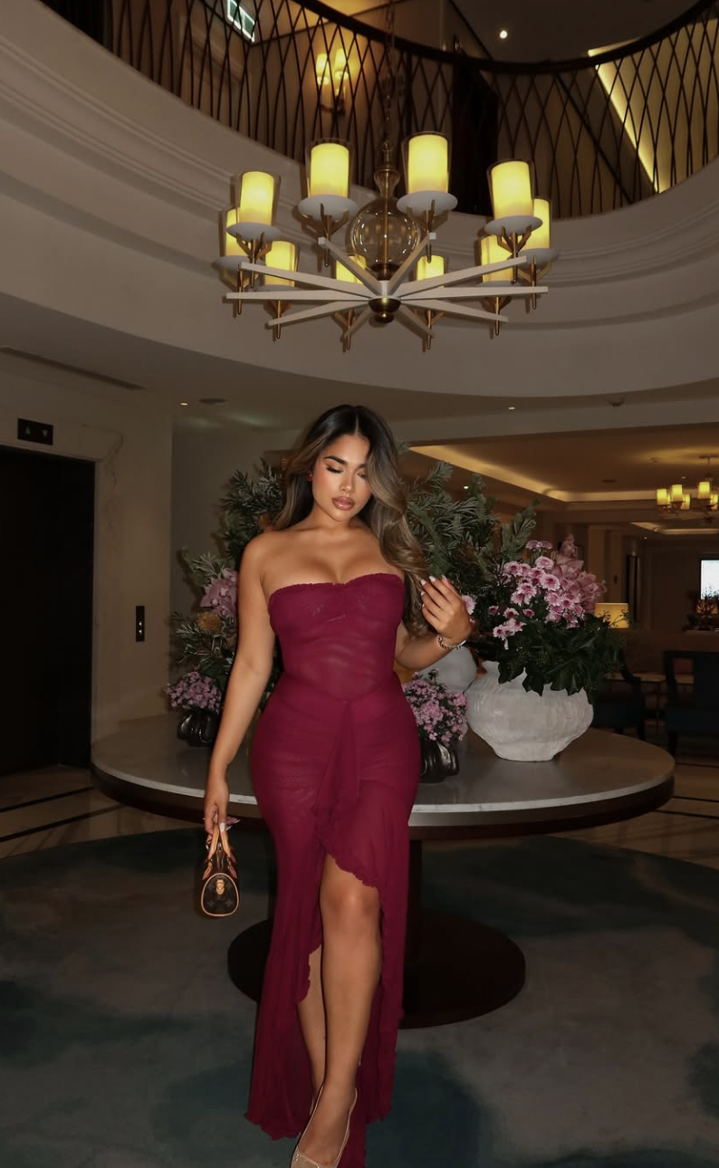 A woman in a red wrap dress with a high leg slit wearing metallic gold strappy high-heeled sandals in a luxury indoor setting.