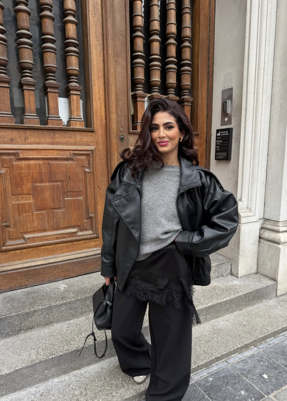 A woman with dark wavy hair wearing a grey knit sweater with a white lace hem, styled under a black oversized leather jacket in front of a wooden architectural door.