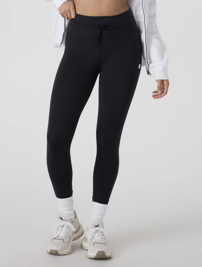 A model wearing the Athleta Rainier High Rise Legging in black, featuring a brushed interior for warmth, a high-waisted fit with a drawcord, and functional side pockets, styled with white crew socks and sneakers.