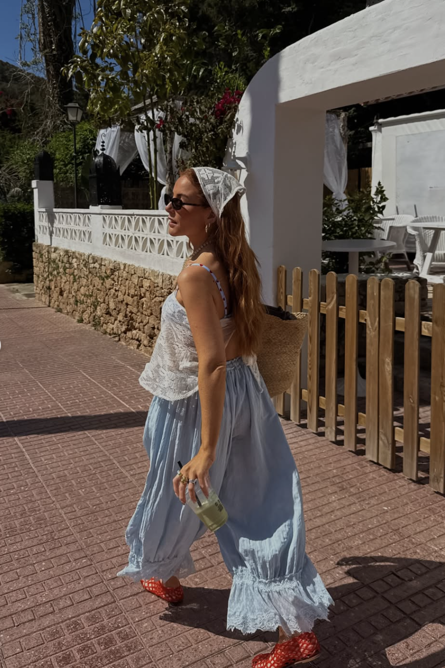 A woman walking outdoors wearing breezy, light blue wide-leg sarong pants with a white lace hem, a white bikini top, a white lace headscarf, and bright red woven lattice jelly flats while holding an iced drink.