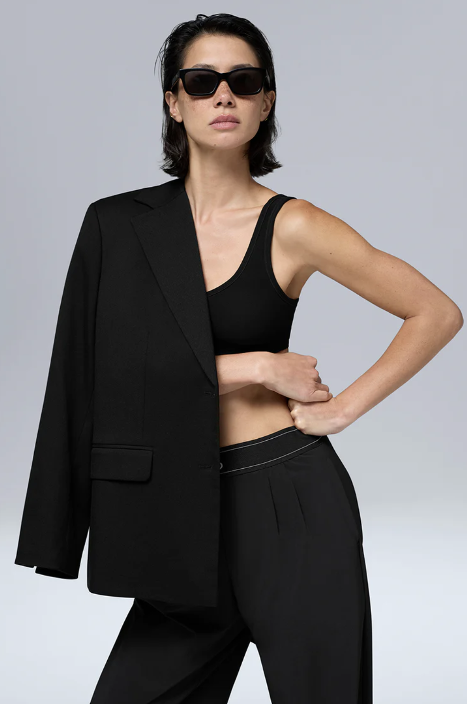 A woman wearing a sleek black blazer over a matching black sports bra tank and black track shorts with a white drawstring detail, accessorized with dark sunglasses for a minimalist street style look.