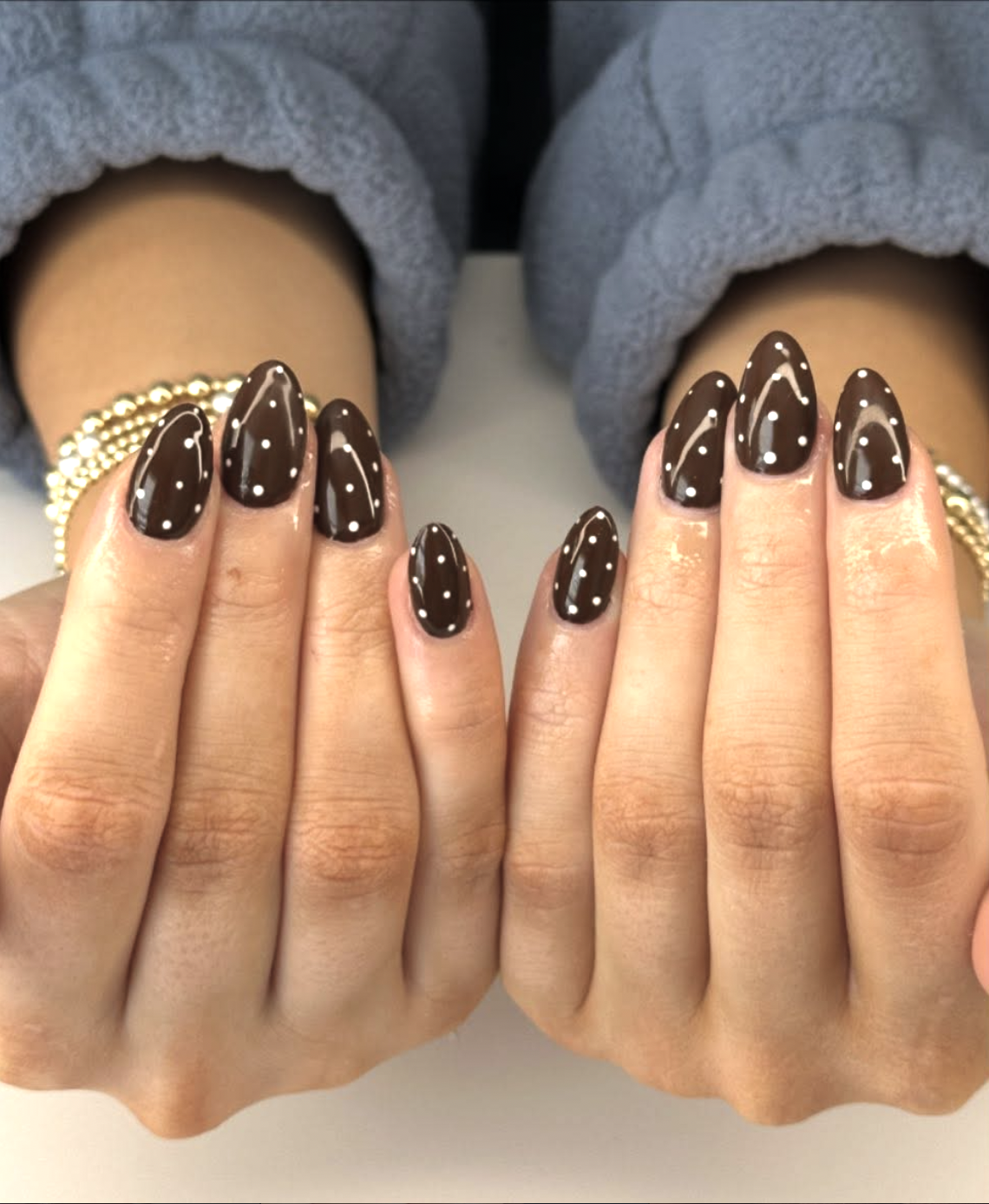 A close-up of almond-shaped nails featuring a dark chocolate brown base with delicate white polka dots, accessorized with gold rings.
