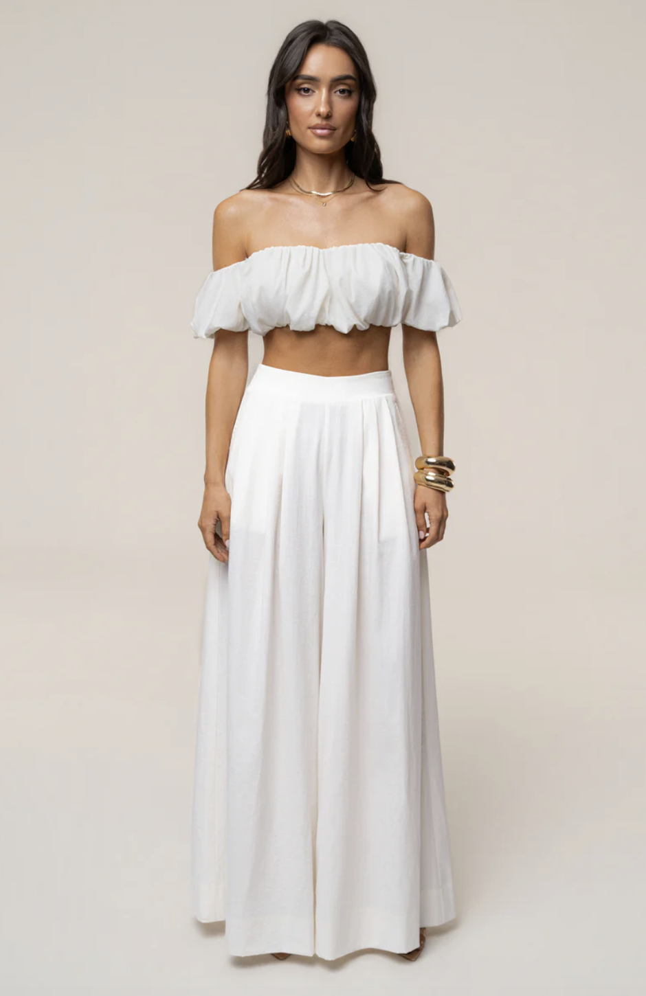 A woman wearing an all-white two-piece set featuring a puffed off-the-shoulder crop top and high-waisted, wide-leg trousers, accessorized with stacked gold cuff bracelets and a delicate gold necklace.