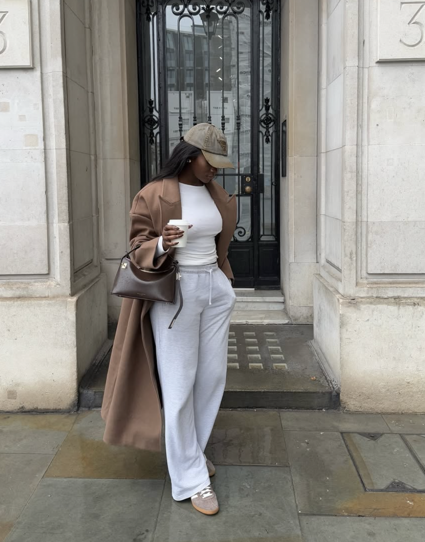 Petite woman wearing a brown wool trench coat, white base layer, and grey joggers paired with tonal low-profile retro sneakers and a tan baseball cap.