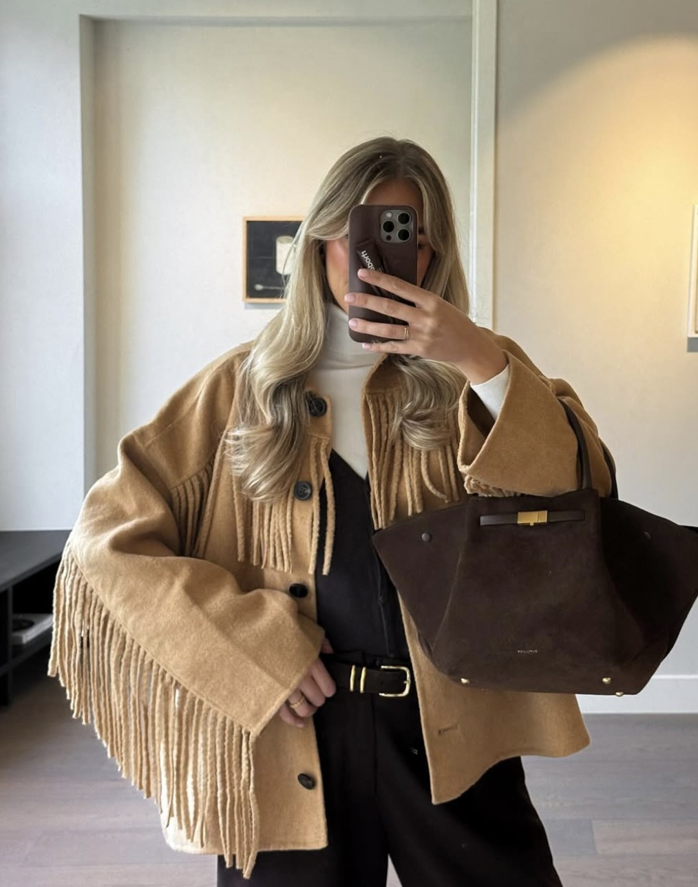 A woman wearing a tan suede fringe jacket over a black outfit and white turtleneck, holding a brown leather bag in a mirror selfie.
