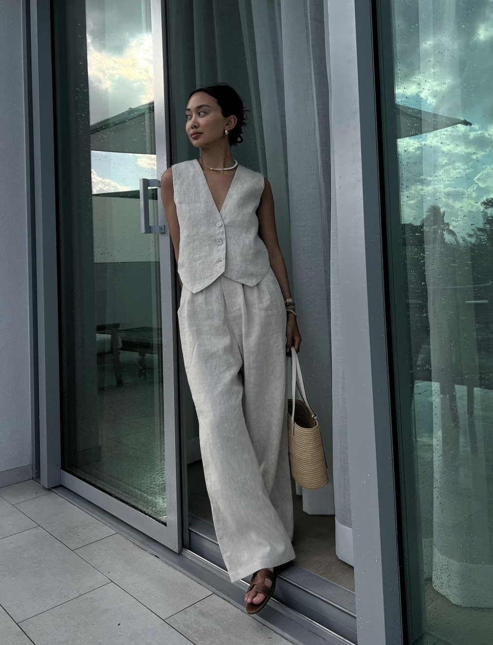 A woman wearing a matching oatmeal-colored linen vest and high-waisted wide-leg linen pants, carrying a straw tote bag and standing by a glass balcony door.