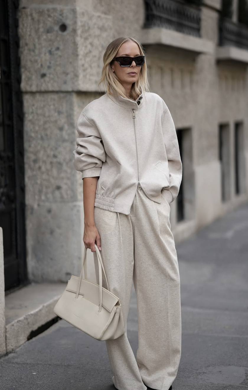 A woman wearing a monochromatic beige leisurewear set consisting of a zip-up sweatshirt and matching joggers, styled with a chocolate brown handbag and dark sneakers.