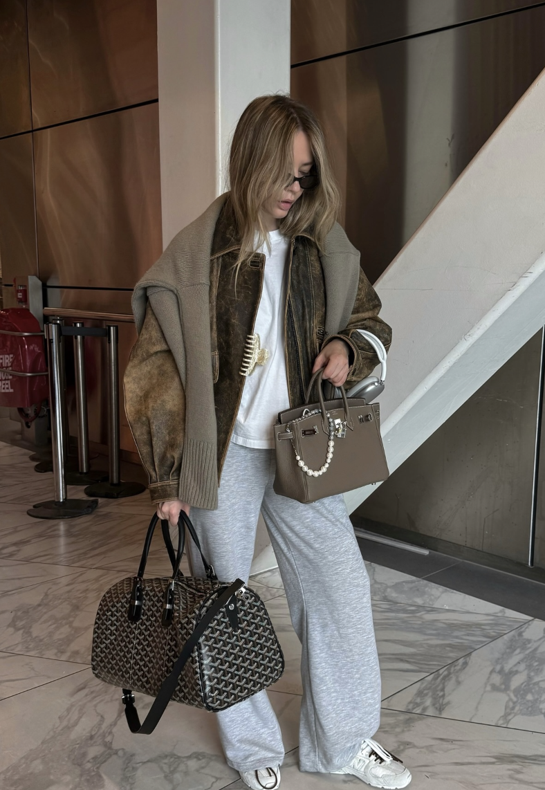 Draped taupe-gray shoulder layer over a white top and gray wide-leg trousers with white sneakers and structured bags.