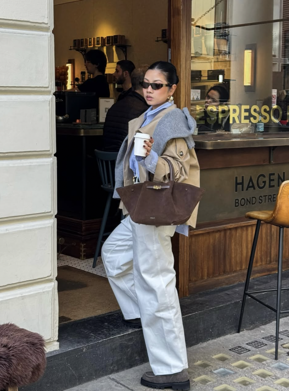 A woman wearing a tan cropped trench coat layered over a blue button-down shirt and grey sweater, paired with wide-leg white trousers and a large brown suede tote bag.